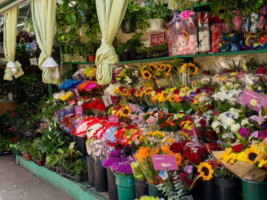 A vibrant outdoor floral display featuring a variety of freshly cut flowers arranged in bouquets and potted plants. The arrangement includes sunflowers with bright yellow petals, white lilies, red and