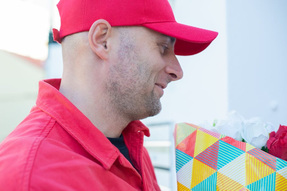 A man dressed in a red cap and matching red jacket is holding a floral gift wrapped in colorful geometric-patterned paper, containing prominent white and red flowers, with green stems and petals visib