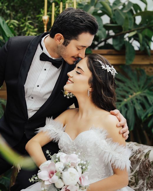 A bride and groom share a tender moment indoors surrounded by lush green tropical plants. The bride, with dark wavy hair adorned with a delicate silver hairpiece, wears an elegant white wedding dress